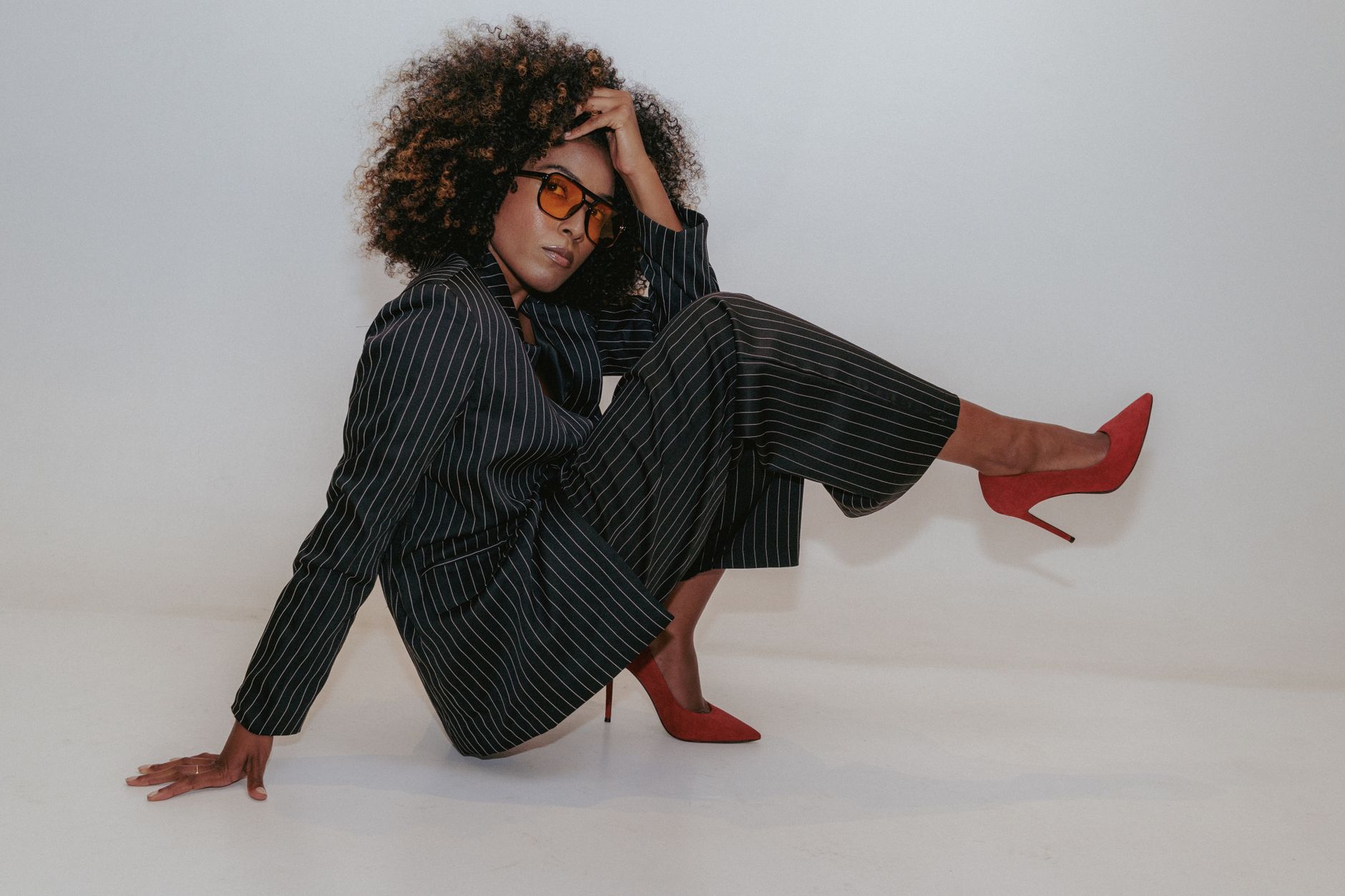 Model in oversized black tailoring and red heels seated on a pale grey studio floor