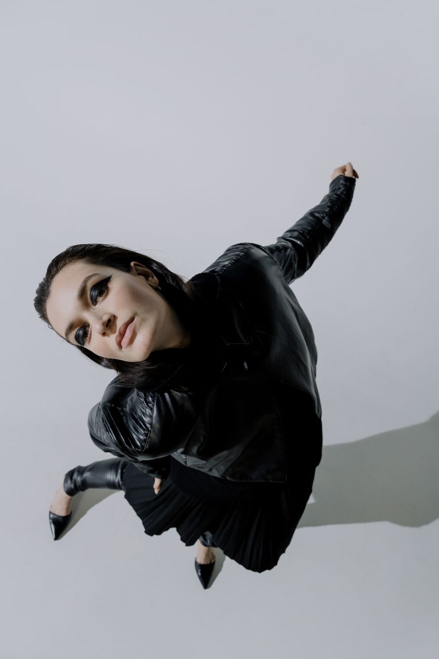 Model in a black leather bomber and pleated skirt lunging toward the camera on a pale studio floor