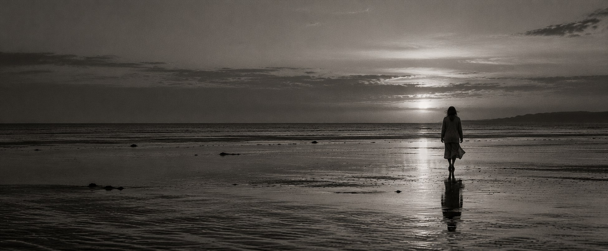 A lone figure walking across wet tidal flats at dusk