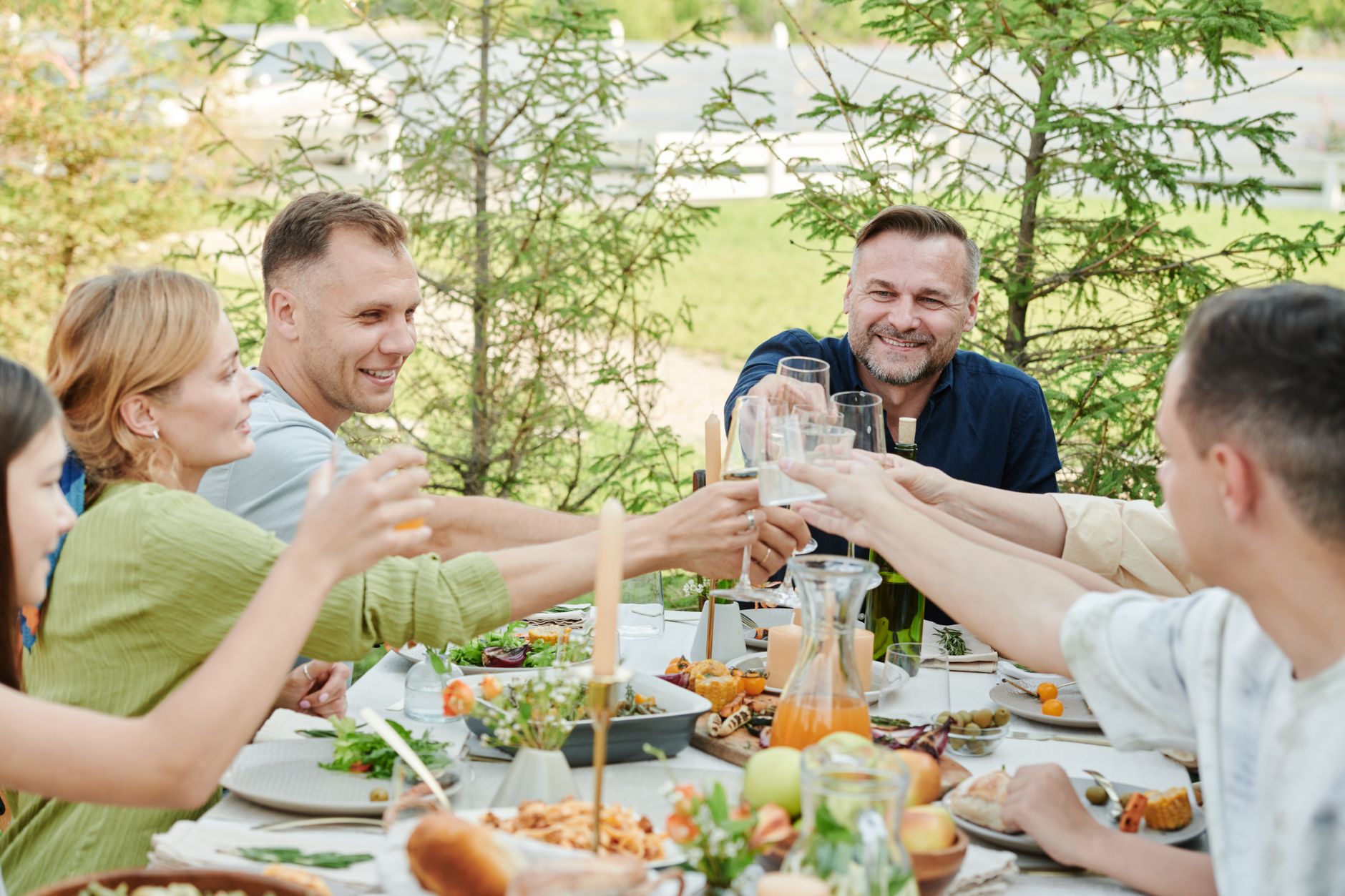 Friends round a garden table in daylight, clinking glasses over salads, bread and a carafe of juice