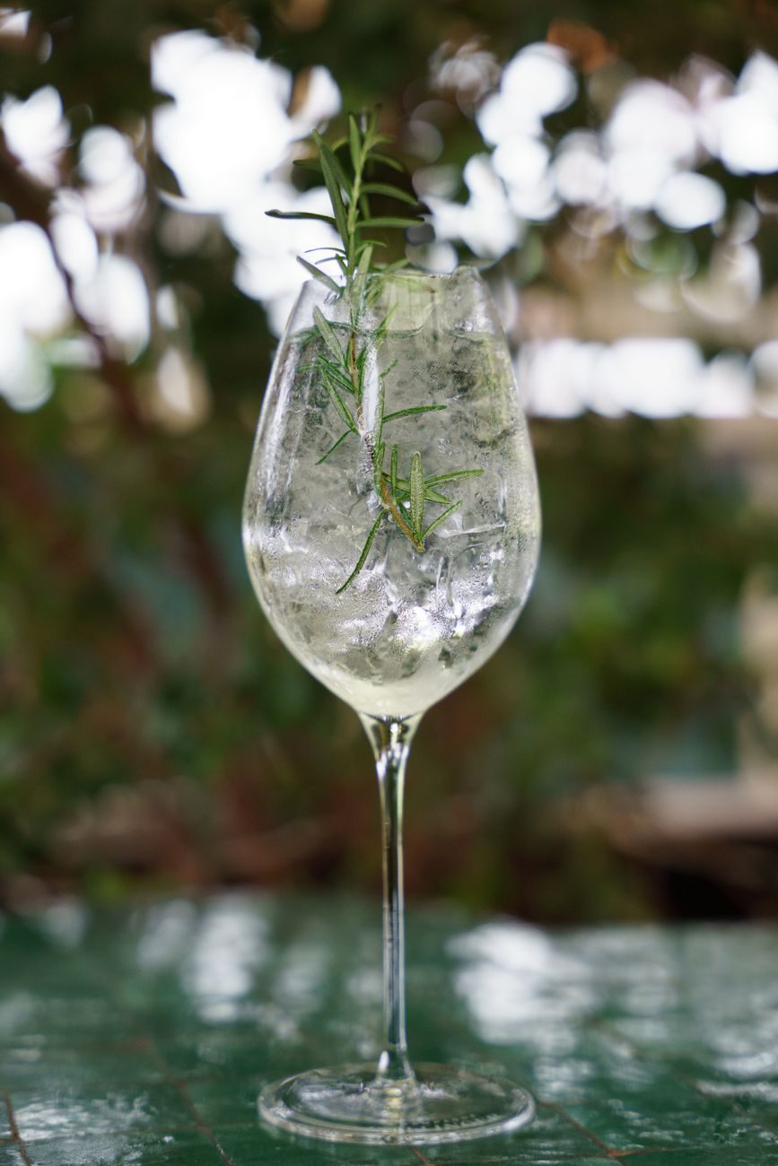 A clear long drink with ice and a rosemary sprig in a stemmed glass, outdoors
