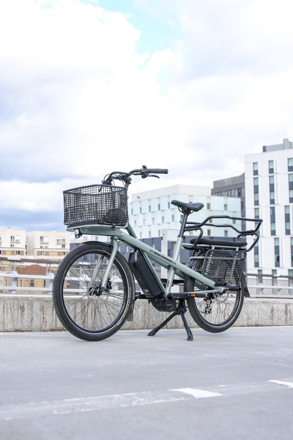 A grey electric cargo bike with a front basket and rear rack parked on a rooftop under a cloudy sky