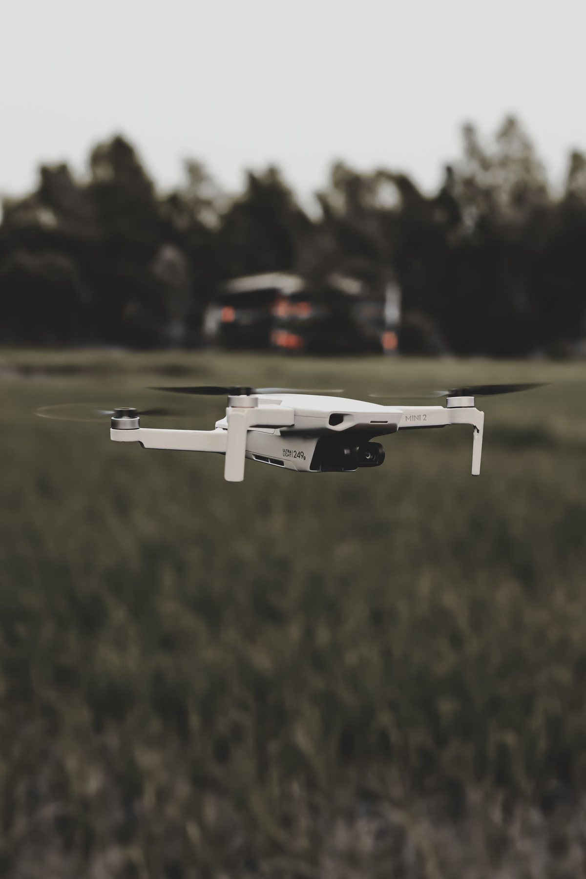 A small white quadcopter drone hovering low over a green field with a tree line behind
