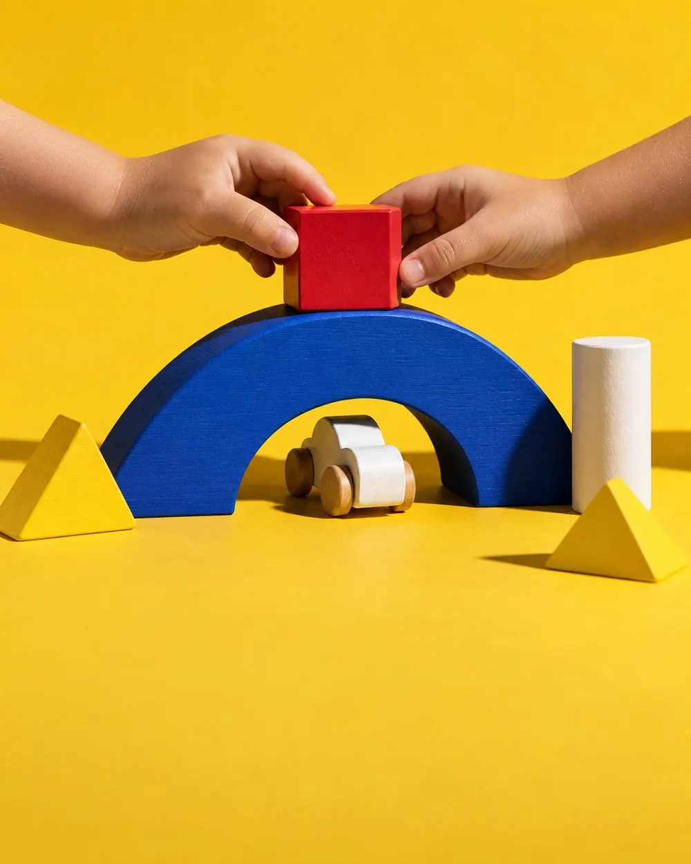 Two children balancing a red cube on a blue arch bridge while a wooden car drives underneath