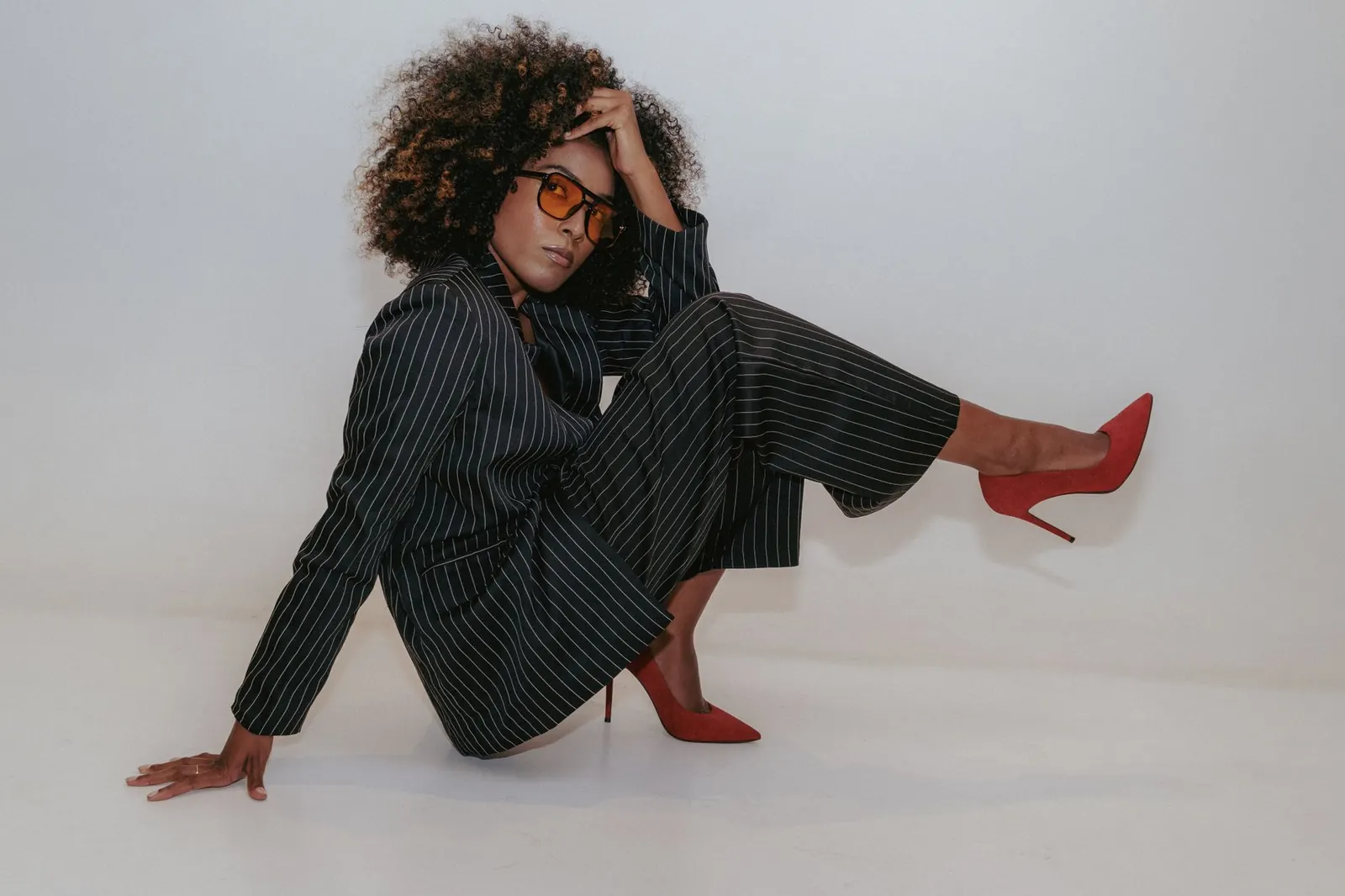 Model in oversized black tailoring and red heels seated on a pale grey studio floor