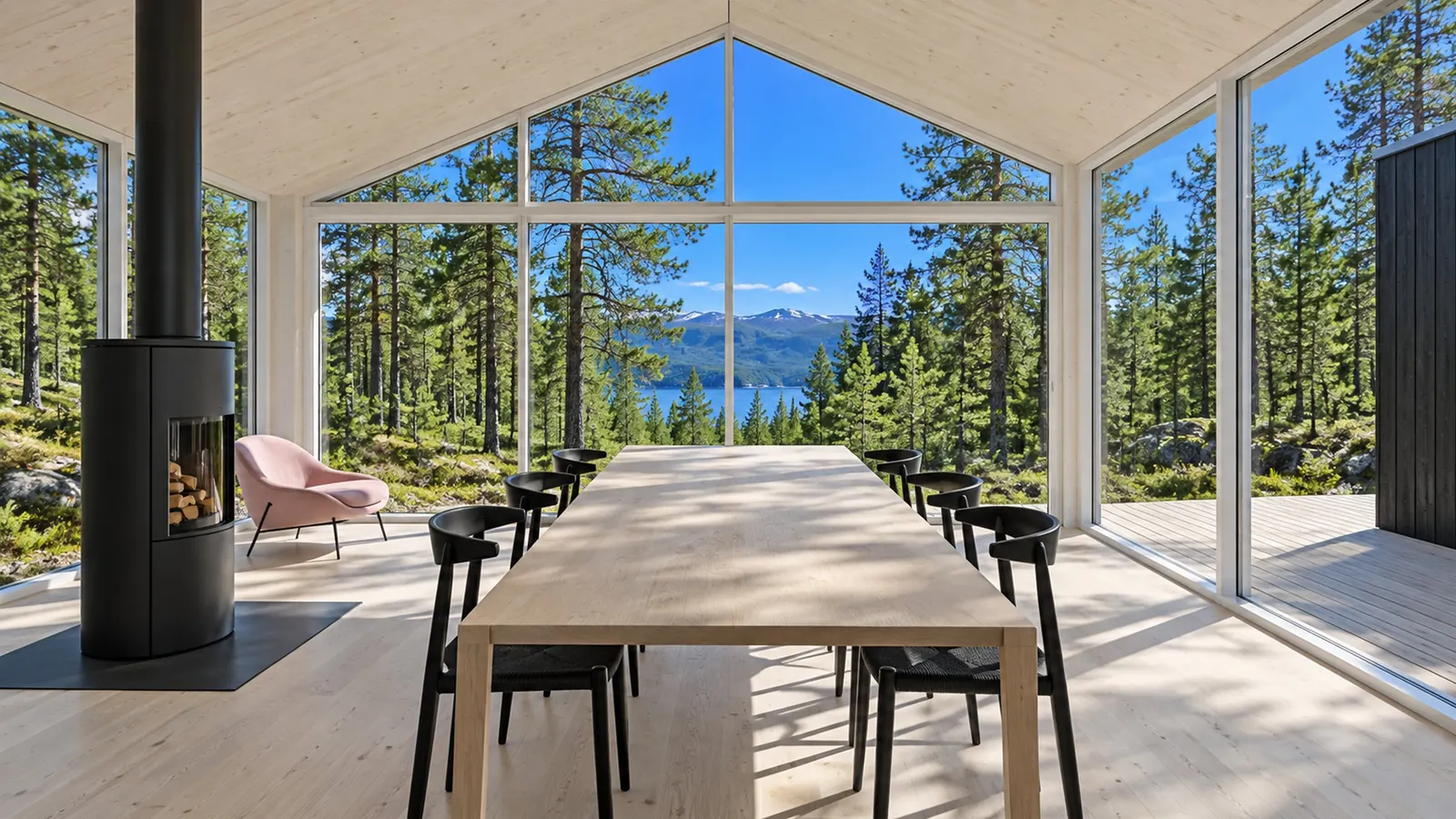 Two-module cabin interior: a long oak table, a black wood stove and a pink chair beside a glass wall onto a pine forest