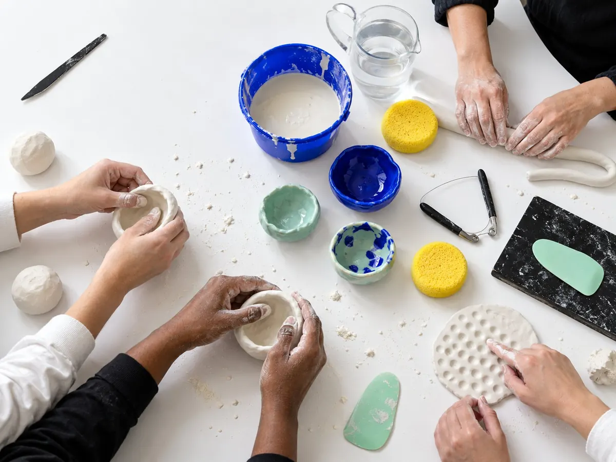 Overhead view of hands pinching small white clay pots around a white table with cobalt and honey tools