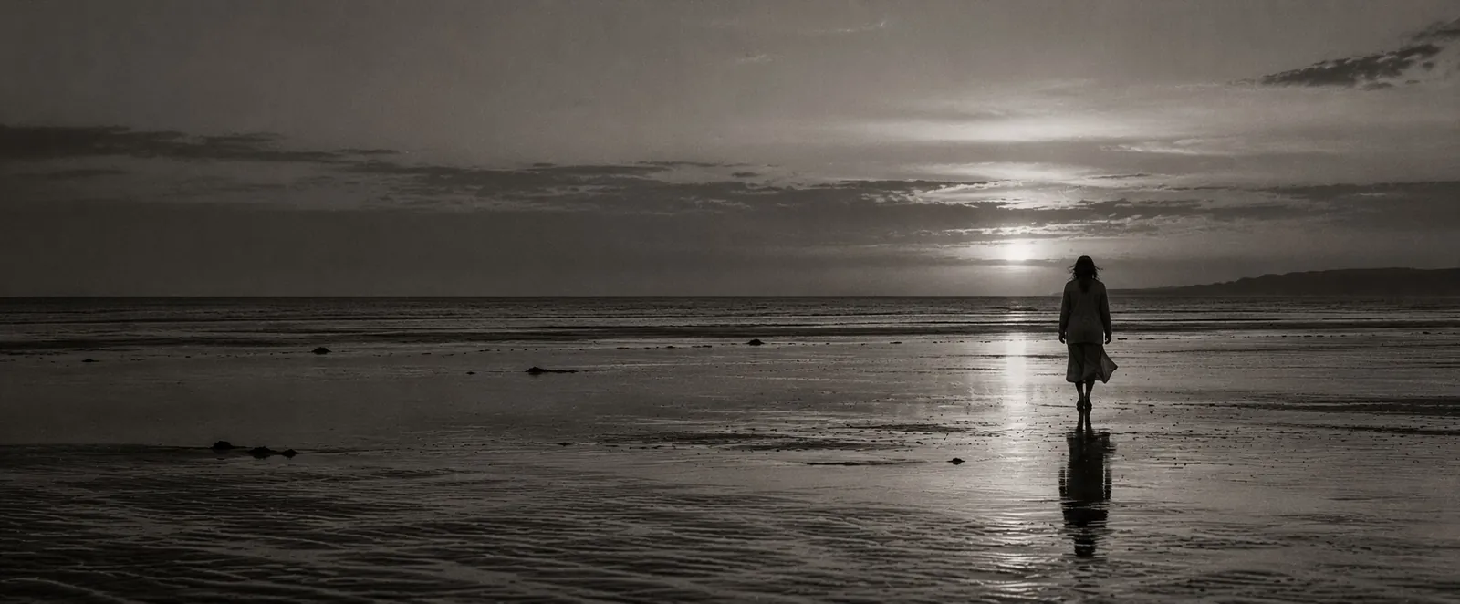 A lone figure walking across wet tidal flats at dusk