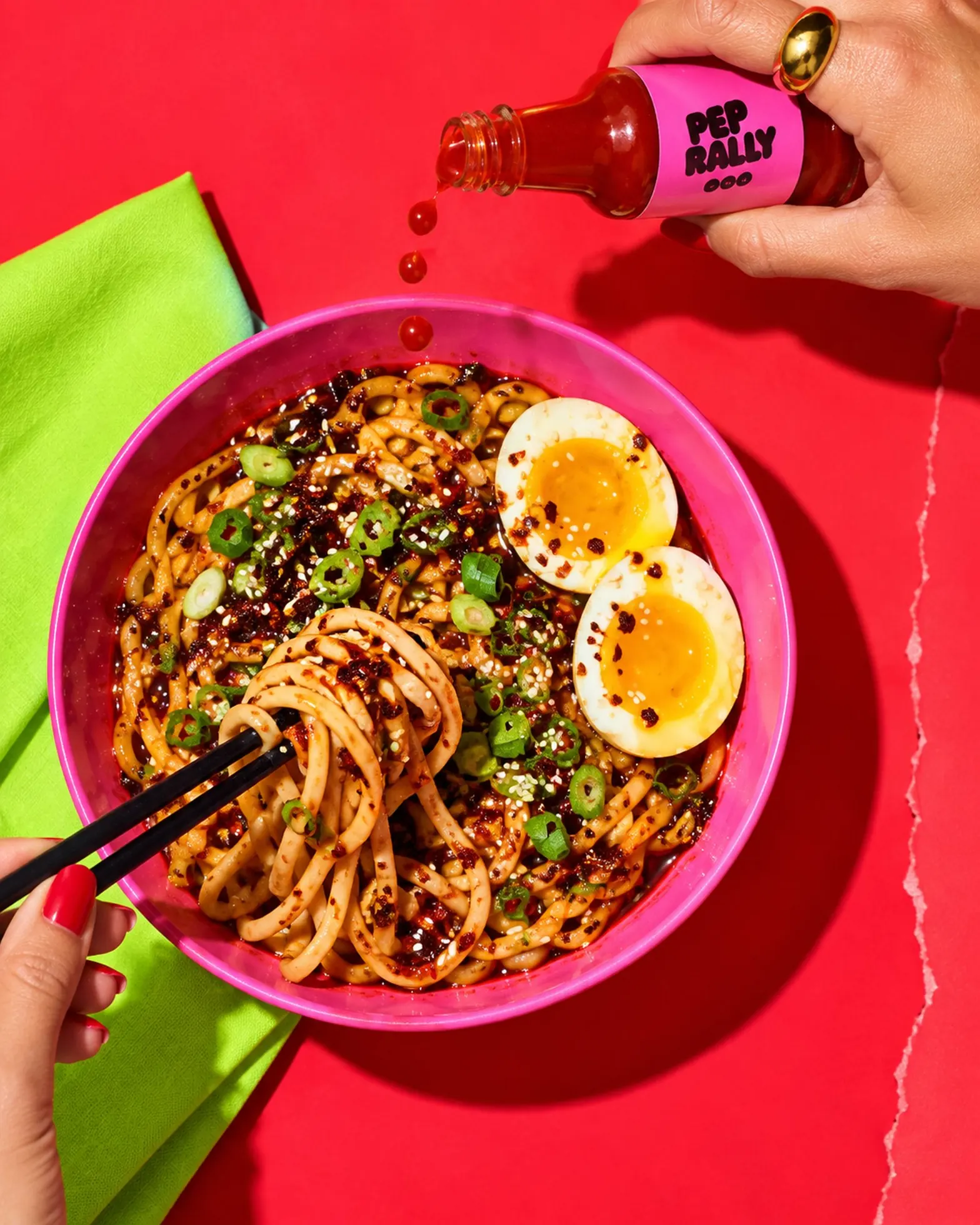 Hands with chopsticks shaking pink-red hot sauce over a bowl of glossy noodles