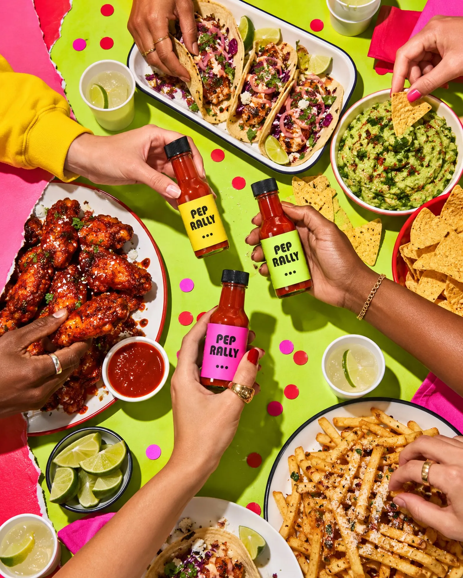 A crowded party table from above with tacos, wings and three hot sauce bottles being passed around