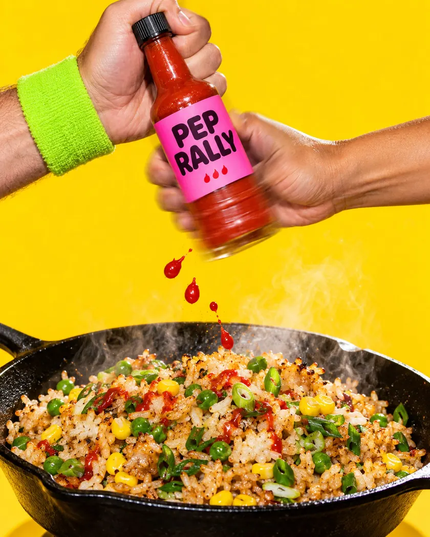 Hands shaking a pink-labelled hot sauce bottle above a sizzling pan