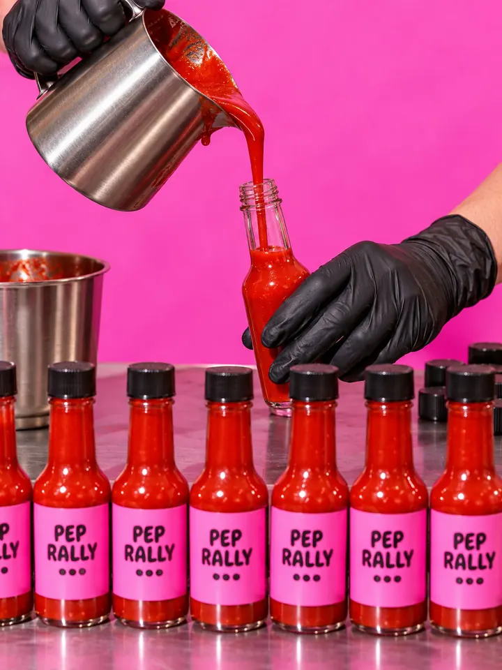 Gloved hands filling small glass bottles with red sauce from a steel jug