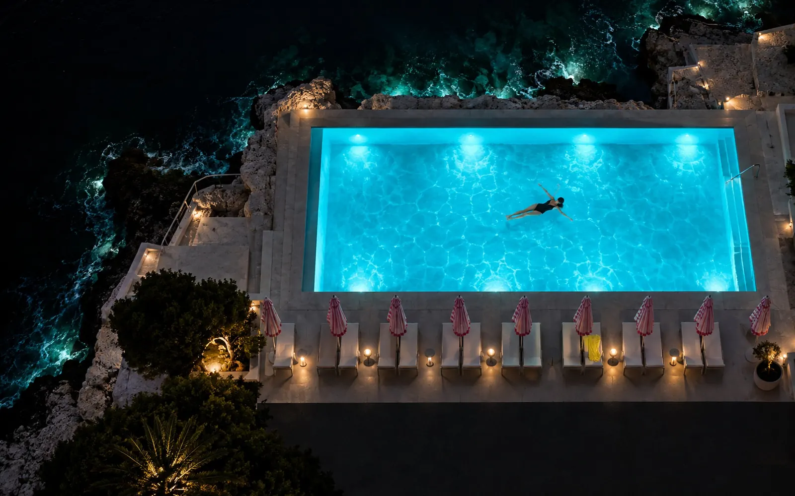 The lido at ten at night from above, the pool glowing blue from its underwater lights and the parasols furled in a row.