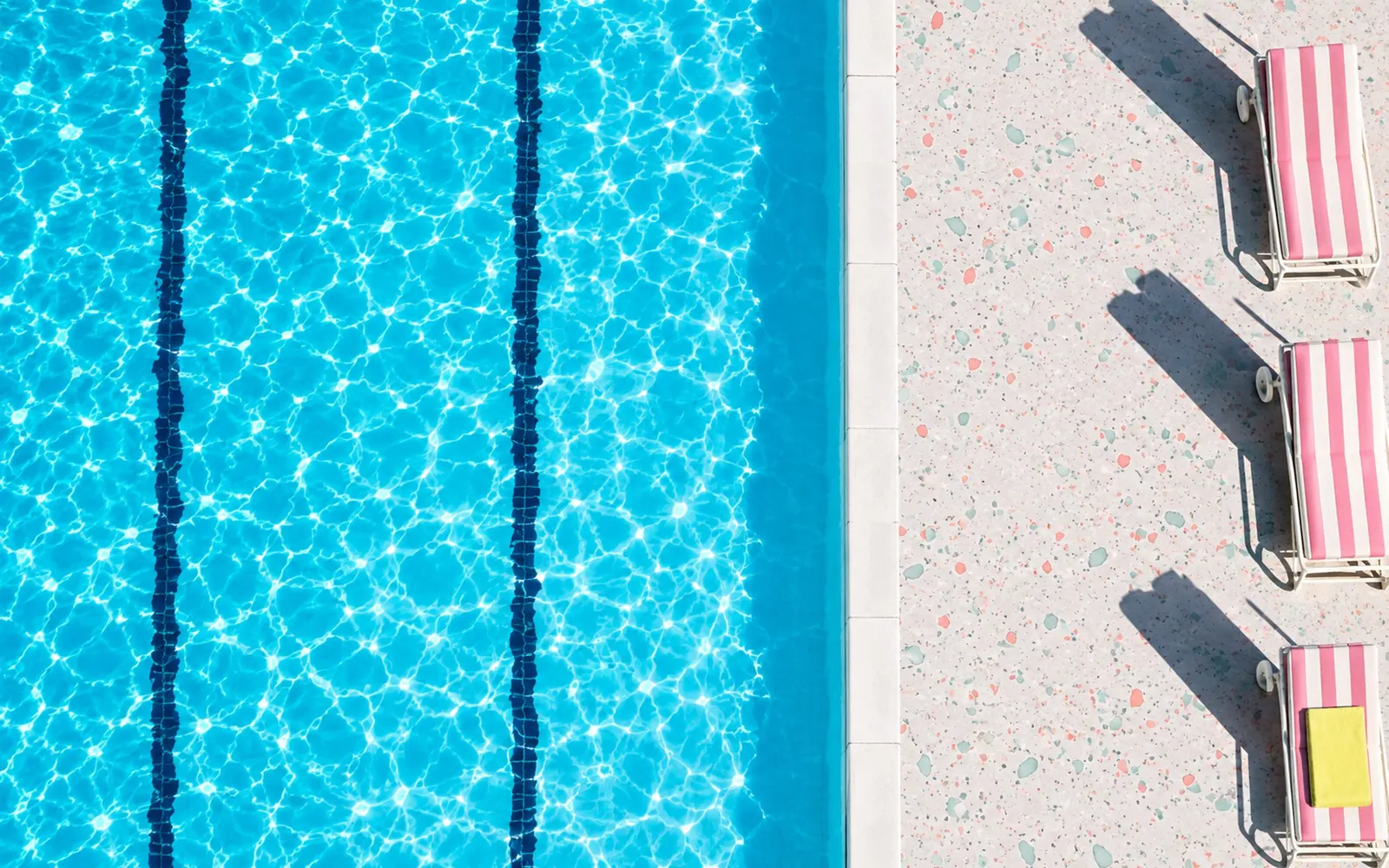 The saltwater lido seen from straight above at seven in the morning: blue water, a white terrazzo deck and folded striped loungers casting long shadows.