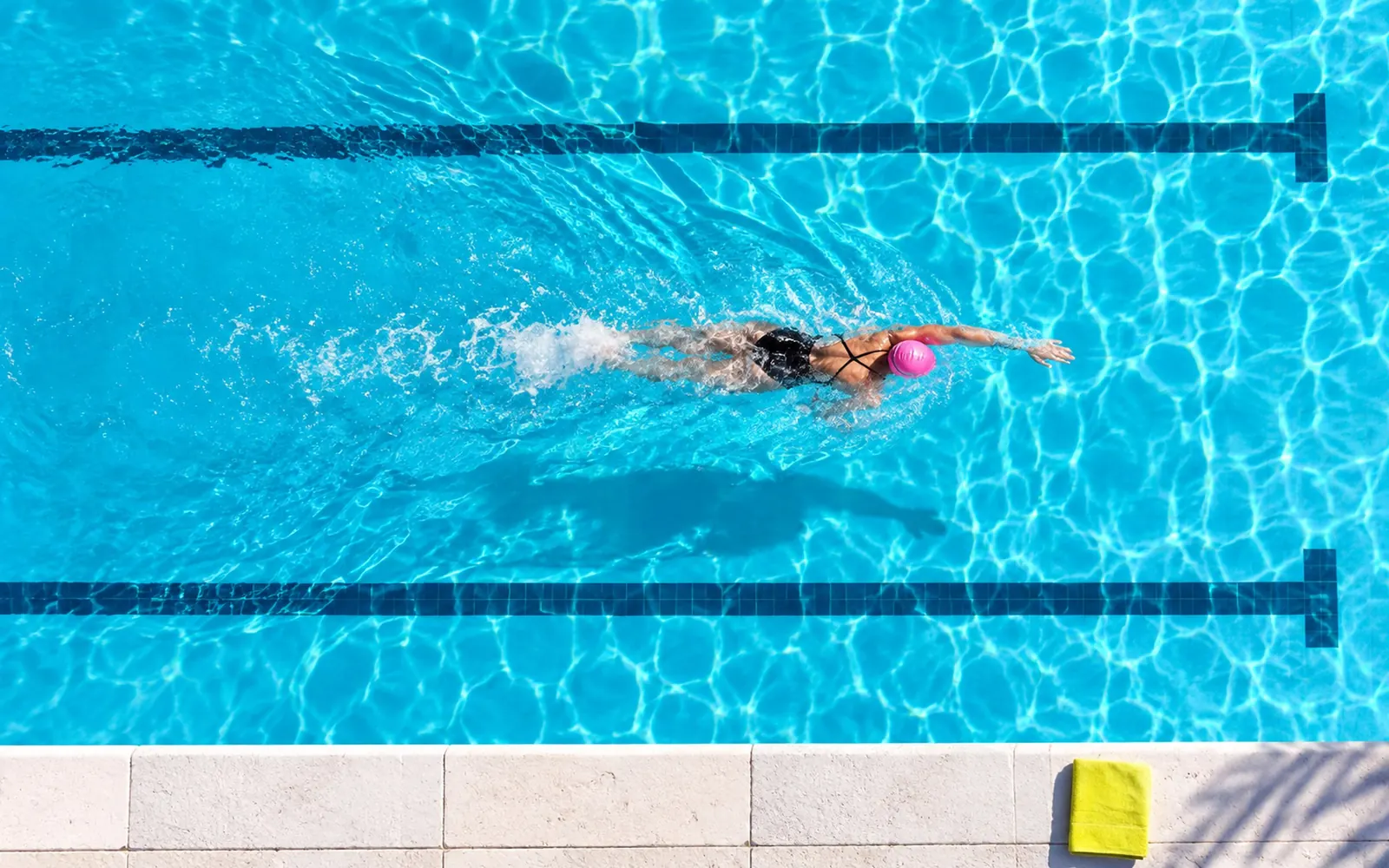 A single swimmer in a pink cap doing front crawl along one lane of the saltwater pool, seen from directly above in low morning light.