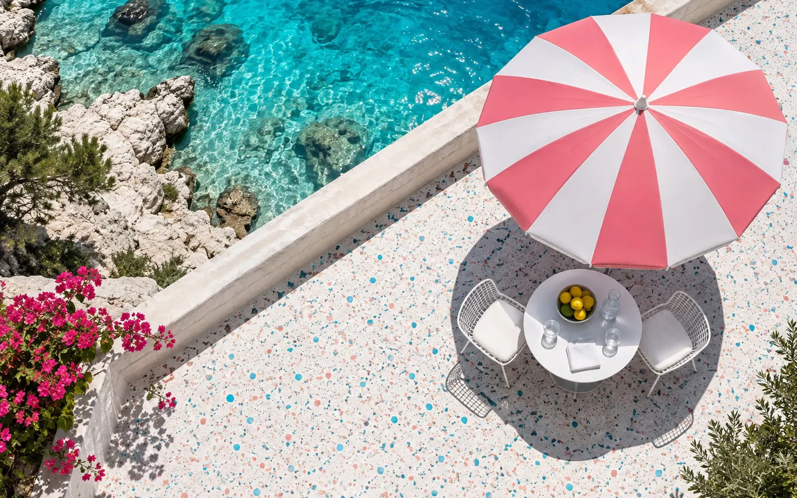 A private terrace from above at noon: a pink and white striped parasol seen as a circle, a white table in its round shadow, the sea beyond a low wall.