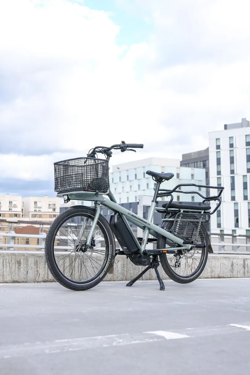 A grey electric cargo bike with a front basket and rear rack parked on a rooftop under a cloudy sky