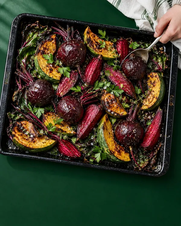 Black roasting tray of blistered beetroot, red carrots and green squash wedges with herbs